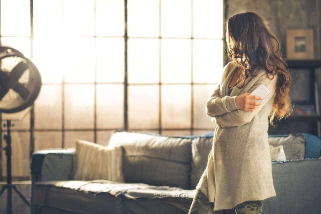 A brunette woman in comfortable clothing is standing in a loft living room holding her phone arms crossed looking away. Urban chic loft decoration details and window.
