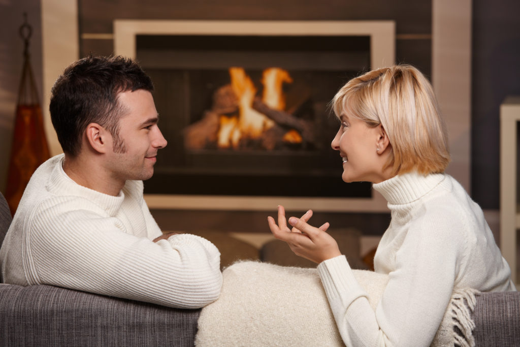 Young romantic couple sitting on sofa in front of fireplace at home, looking at each other, talking.